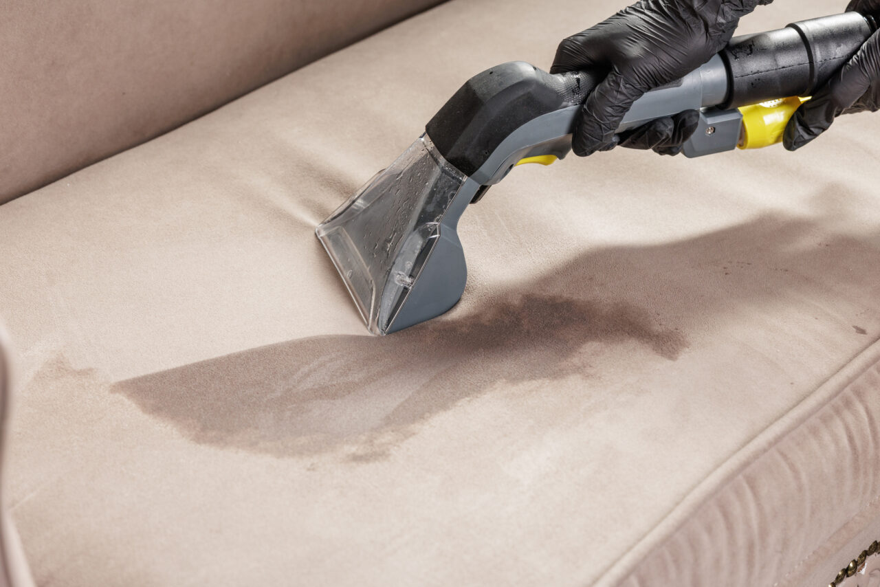 Close-up of a gloved worker using an upholstery extractor on a wet sofa stain, ideal for a cleaning company website banner or brochure about stain removal.